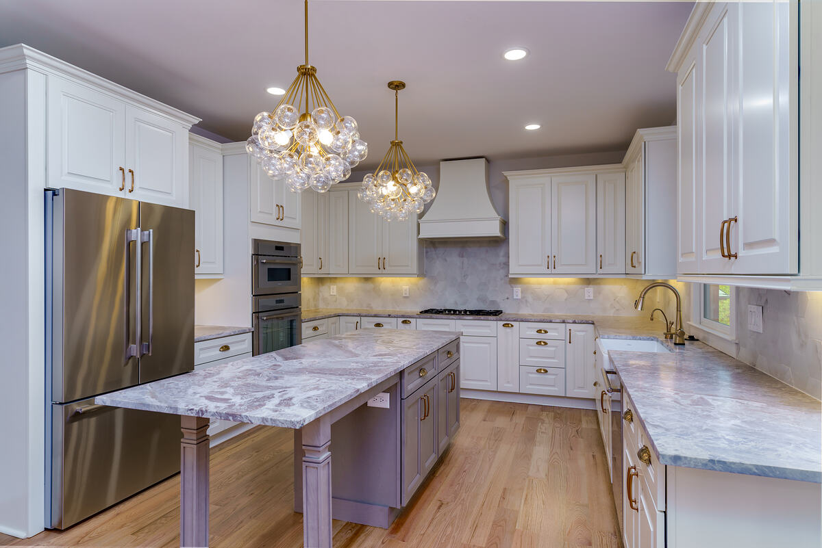 A white kitchen with a marble island, brass pendants, and paneled appliances