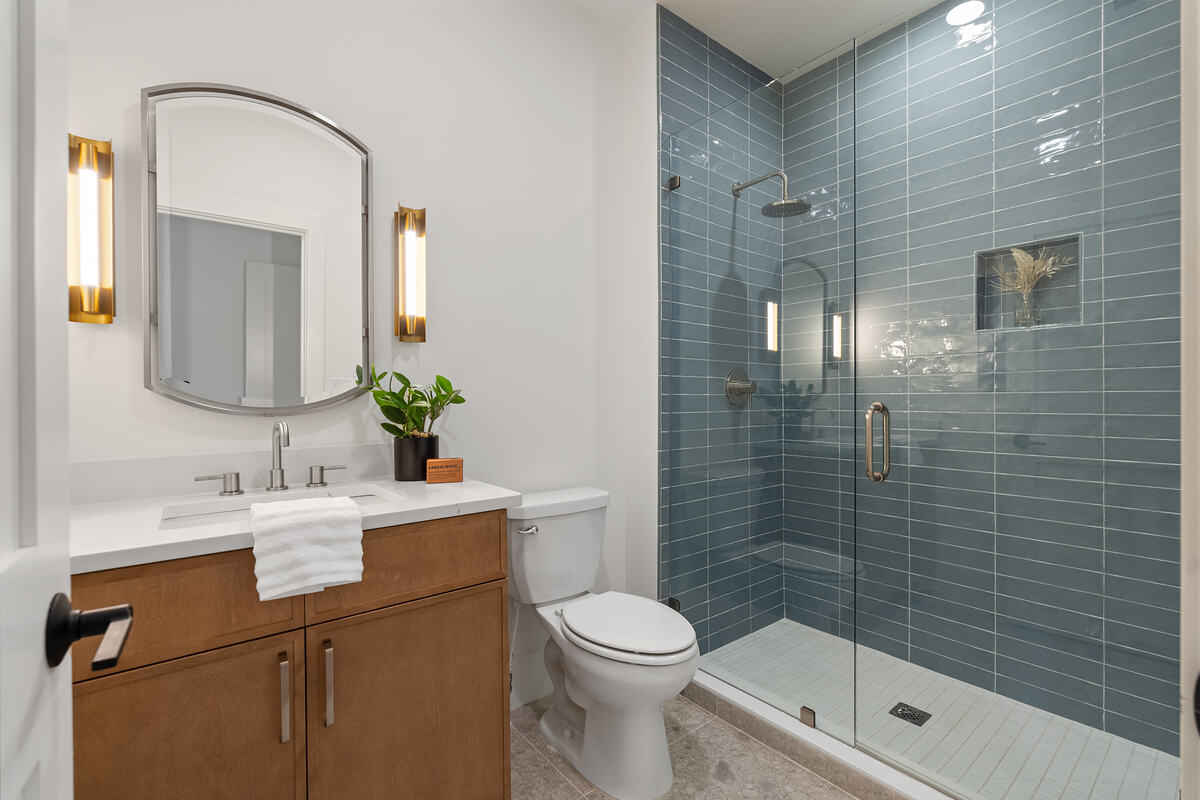 A guest bathroom with blue-green tiled shower, wood vanity, and brass sconces